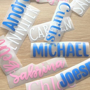 May include: A collection of adhesive vinyl name decals in various fonts and colors, including blue, pink, and white. The names visible include Andre, Jennifer, Curtis, Carmen, Michael, Beth, Sabrina, Joseph, and Chloe, printed on clear backing paper.