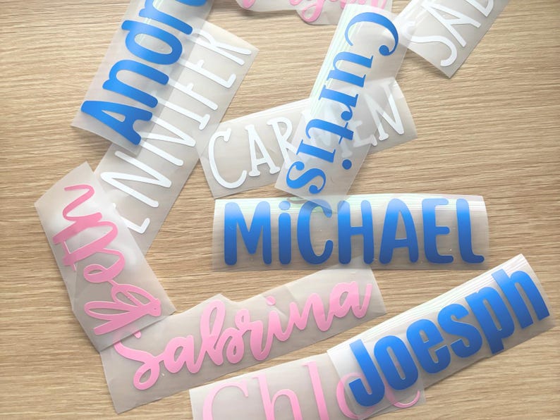 May include: A collection of adhesive vinyl name decals in various fonts and colors, including blue, pink, and white. The names visible include Andre, Jennifer, Curtis, Carmen, Michael, Beth, Sabrina, Joseph, and Chloe, printed on clear backing paper.