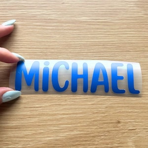 May include: A blue vinyl decal featuring the name MICHAEL in a bold, sans-serif font. The letters are cut from a solid blue adhesive material and are presented on a clear transfer backing, ready for application onto a smooth surface.