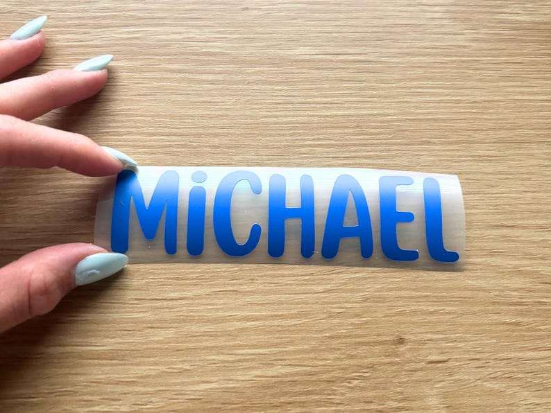 May include: A blue vinyl decal featuring the name MICHAEL in a bold, sans-serif font. The letters are cut from a solid blue adhesive material and are presented on a clear transfer backing, ready for application onto a smooth surface.