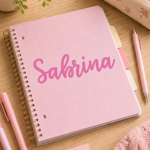 May include: A light pink spiral-bound notebook featuring the name Sabrina printed in a dark pink script font on the front cover. The notebook includes several colored tabbed dividers along the side and is bound with a gold-toned metal coil.