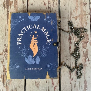 May include: A navy blue book with the words "PRACTICAL MAGIC" in white, featuring a hand, stars, and crystals design. The book has a gold border, a black leather clasp, and a silver chain. The author's name, Alice Hoffman, is printed below.