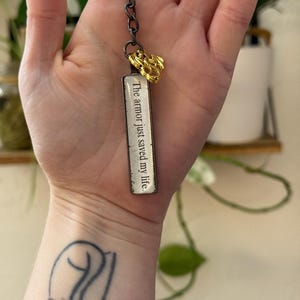 Puede incluir: Un llavero plateado con un pequeño amuleto de dragón dorado y una etiqueta rectangular que dice "The armor just saved my life."