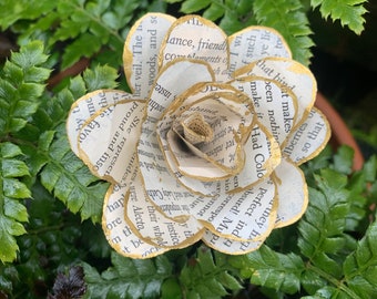 Puede incluir: Una rosa de papel hecha a mano con bordes dorados, elaborada con páginas de libros. La flor está rodeada de hojas de helecho verdes, creando una exhibición natural y artística. Los pétalos muestran texto.