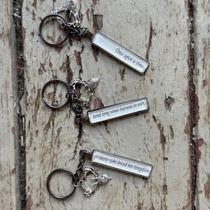 Puede incluir: Tres llaveros plateados con colgantes rectangulares. Cada colgante tiene un fondo blanco con texto negro: "Once upon a time," "land long since burned to ash," y "princess who loved her kingdom."