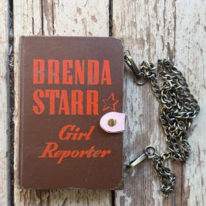 以下が含まれることがあります： オレンジ色の「BRENDA STARR Girl Reporter」の文字が入った、茶色の本型バッグ。 ピンク色の留め金とシルバーのチェーンストラップが付いています。 背景は風化した木製の表面です。