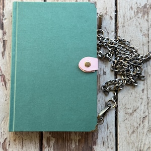 May include: A teal-colored journal with a pink clasp and a metal chain. The journal has gold-colored corner protectors and a vertical gold stripe on the spine. The chain is a dark silver color.