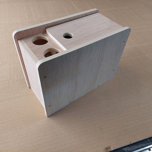 May include: A wooden box with a hinged lid and two round holes on the front. The box is made of light-coloured wood and has a natural finish.