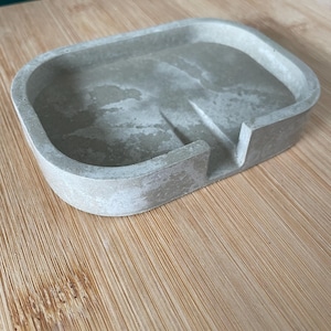 Könnte beinhalten: Ein rechteckiger, hellgrauer Seifenhalter aus Beton mit abgerundeten Kanten. Der Halter hat einen erhöhten Rand und einen kleinen, abgewinkelten Abschnitt zur Entwässerung. Die Oberfläche hat ein gesprenkeltes Aussehen. Der Halter steht auf einer hellbraunen Holzoberfläche.