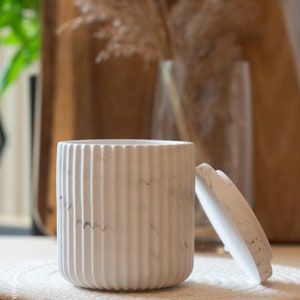 Könnte beinhalten: Weiße Keramikdose mit Deckel, mit Marmorierung und vertikalen Rillen. Die Dose steht auf einer beigefarbenen gewebten Matte.