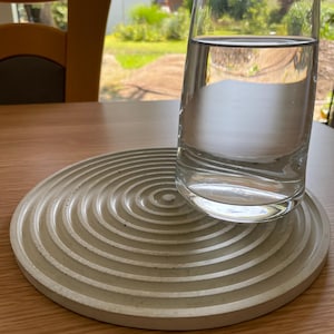 Op de afbeelding: Een heldere glazen waterkan staat op een cirkelvormige, lichtgrijze betonnen onderzetter met concentrische groeven. De onderzetter staat op een lichtbruine houten tafel.