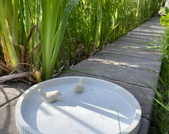 May include: A light gray, round concrete bird bath with a ribbed edge, featuring two small white bird figurines. The bird bath is placed on a wooden walkway, surrounded by lush green plants and grass.