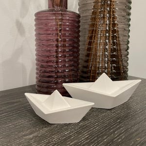 Könnte beinhalten: Zwei weiße, papierbootförmige Dekorationen stehen auf einer dunklen Oberfläche. Dahinter befinden sich zwei Glasvasen, eine lila und eine braune, beide mit geripptem Design. Die Vasen sind leer.