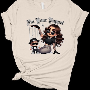 May include: A beige t-shirt featuring a graphic of a woman holding a puppet. The woman has long brown hair, black sunglasses, and a black and white outfit. The puppet is a man with a black hat, sunglasses, and a black and white outfit. The text "I'm Your Puppet" is written in black above the graphic.