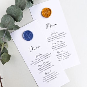 May include: Two white cards with a menu for a wedding or special event. The cards are decorated with wax seals, one blue and one gold. The menu lists a starter, main course, and dessert. The starter is grilled fillet with red onion, served with baby potatoes, carrots, green beans and curly kale. The main course is herb roasted lamb and rice with fresh basil, spring vegetable risotto. The dessert is dark chocolate cake, selection of ice creams, sticky toffee pudding with vanilla ice cream.