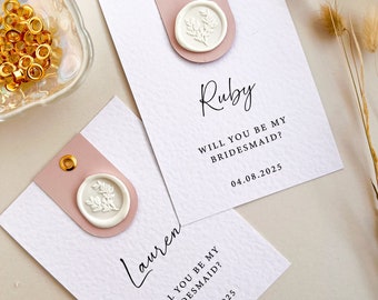 May include: Two white cards with pink accents and gold eyelets. Each card has a wax seal with a floral design and the text "Will you be my bridesmaid?" with a date. The names "Lauren" and "Ruby" are written in cursive on the cards.