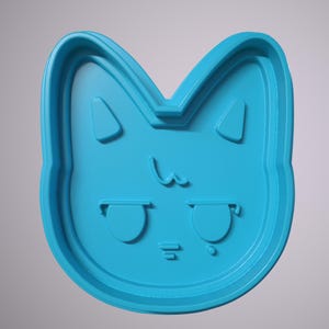 May include: A blue plastic cookie cutter shaped like a cat's face with a cute expression. The cat has large eyes, a small nose, and a small mouth.