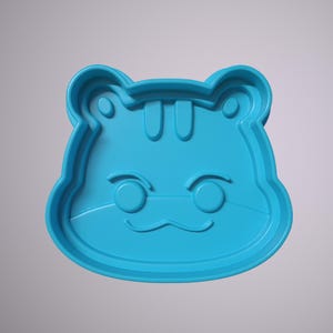 May include: A blue plastic cookie cutter shaped like an animal with a cute face. The cutter has a rounded shape with ears, eyes, and a mouth.