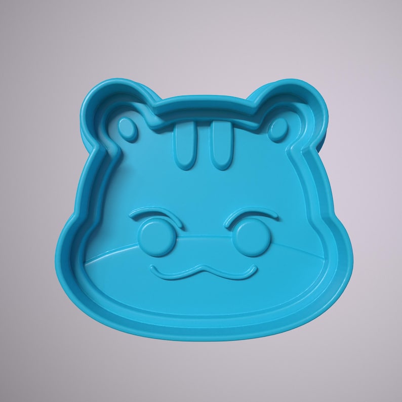 May include: A blue plastic cookie cutter shaped like an animal with a cute face. The cutter has a rounded shape with ears, eyes, and a mouth.