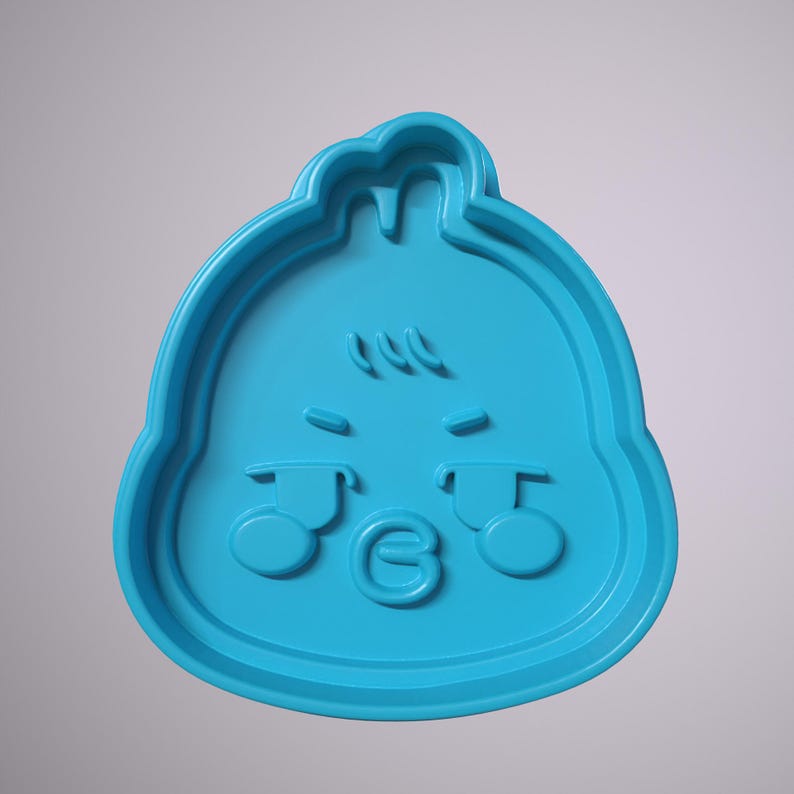 May include: Blue plastic cookie cutter shaped like a cartoon face with a grumpy expression. The face has two large eyes, a small mouth, and a few hairs on top.