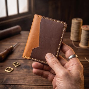 Puede incluir: Una cartera de cuero plegable con un diseño de dos tonos, que presenta un cuerpo principal de color marrón oscuro y un panel de acento de cuero color canela más claro en el lado izquierdo. La cartera tiene costuras amarillas visibles.