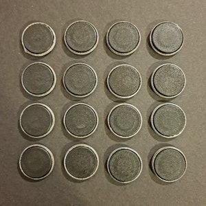 May include: Sixteen round, black magnets with silver-colored metal rims are arranged in a square grid on a dark surface. The magnets have a flat, matte finish and are evenly spaced.
