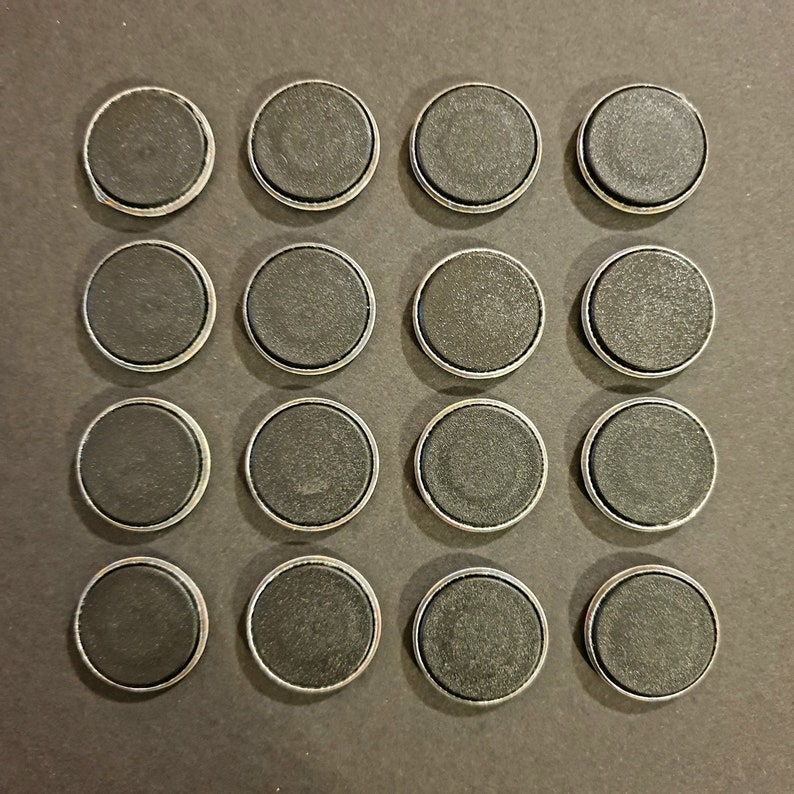 May include: Sixteen round, black magnets with silver-colored metal rims are arranged in a square grid on a dark surface. The magnets have a flat, matte finish and are evenly spaced.