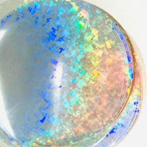 May include: A stack of clear, iridescent, circular stickers. The stickers have a holographic effect with a pattern of small, colorful squares. The colors include blue, green, yellow, and pink, creating a shimmering appearance.