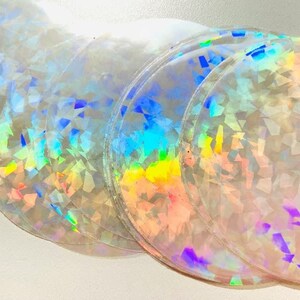 May include: A stack of iridescent, circular stickers. The stickers have a clear base with a holographic, geometric pattern that reflects a spectrum of colors, including blue, green, yellow, and pink. The edges of the stickers are slightly raised.