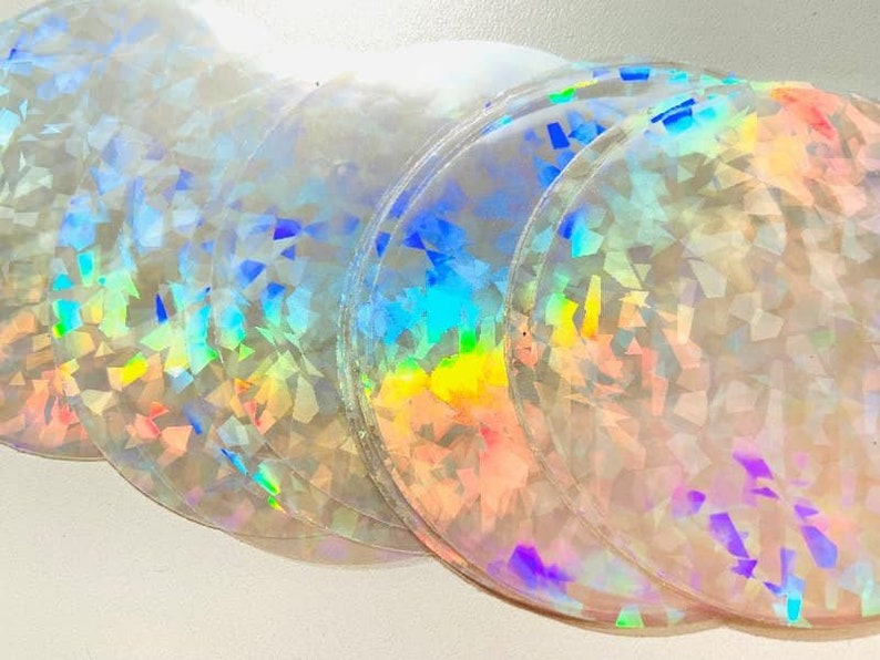 May include: A stack of iridescent, circular stickers. The stickers have a clear base with a holographic, geometric pattern that reflects a spectrum of colors, including blue, green, yellow, and pink. The edges of the stickers are slightly raised.