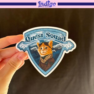 May include: A sticker featuring a cartoon cat wearing aviator goggles and a brown jacket. The cat is set against a blue sky and cloud background, with the words "Quest Squad" in a banner above. The sticker has a white border.
