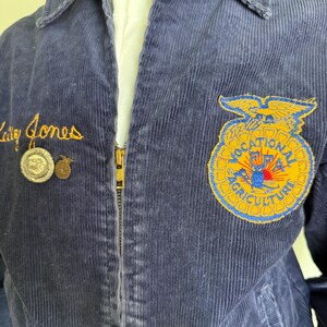 Op de afbeelding: Marineblauwe corduroy jas met een gouden rits en geborduurde details. Voorzien van een patch met de woorden "VOCATIONAL FFA AGRICULTURE" en de naam "Cathy Jones" in gouden script. Inclusief twee kleine spelden.
