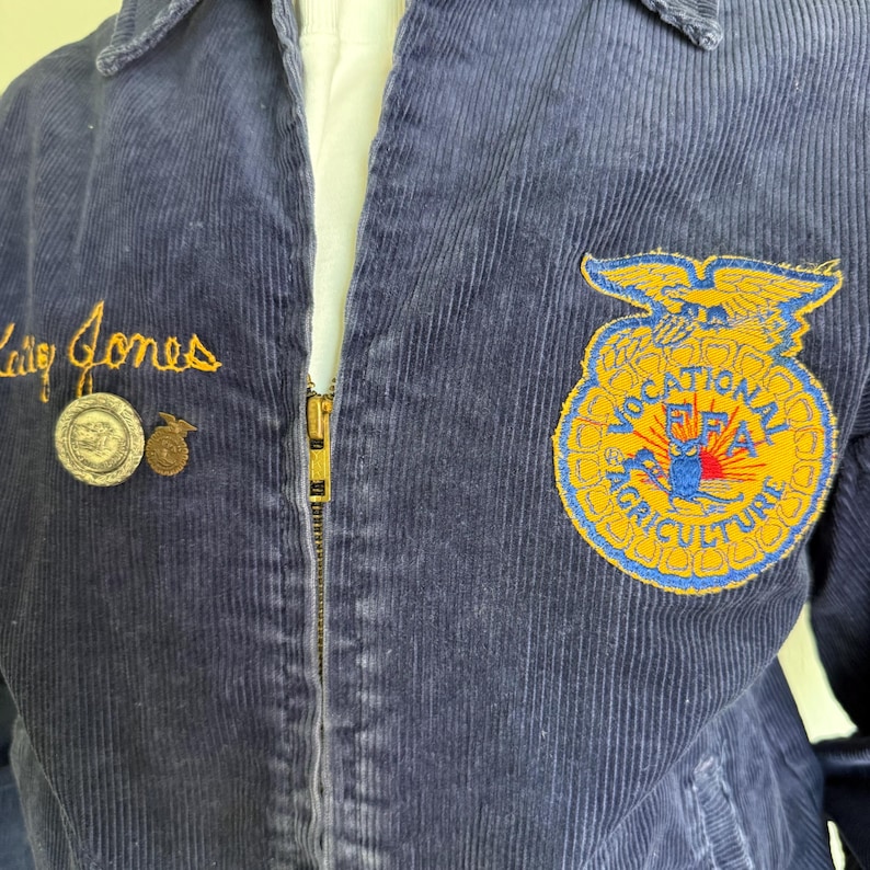 Op de afbeelding: Marineblauwe corduroy jas met een gouden rits en geborduurde details. Voorzien van een patch met de woorden "VOCATIONAL FFA AGRICULTURE" en de naam "Cathy Jones" in gouden script. Inclusief twee kleine spelden.