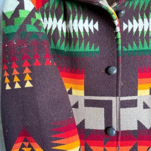 Puede incluir: Primer plano de un abrigo abotonado con un patr&oacute;n geom&eacute;trico colorido. El abrigo presenta una base marr&oacute;n con vibrantes acentos de rojo, naranja, amarillo, verde y blanco. El abrigo tiene tres botones de color gris oscuro.