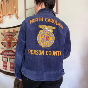 Op de afbeelding: Marineblauwe corduroy jas met de woorden "NORTH CAROLINA" en "PERSON COUNTY" in het geel geborduurd. Een cirkelvormig embleem met een adelaar en de letters "FFA" is ook op de achterkant van de jas geborduurd.