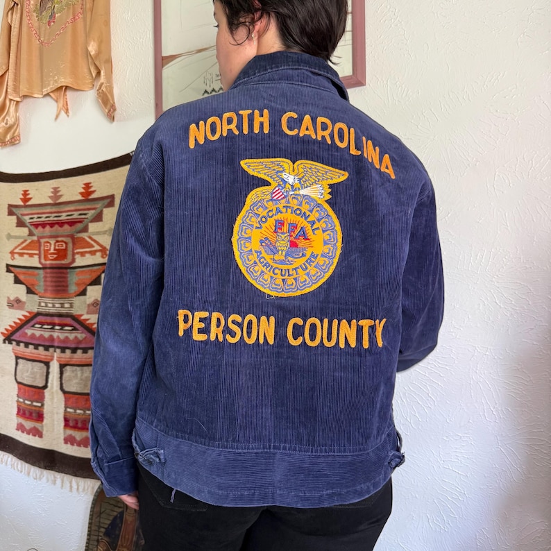 Op de afbeelding: Marineblauwe corduroy jas met de woorden "NORTH CAROLINA" en "PERSON COUNTY" in het geel geborduurd. Een cirkelvormig embleem met een adelaar en de letters "FFA" is ook op de achterkant van de jas geborduurd.