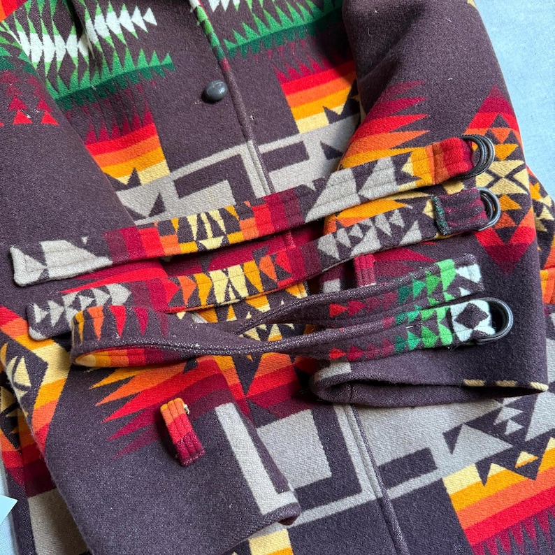 Puede incluir: Primer plano de un abrigo de lana marr&oacute;n con un colorido estampado geom&eacute;trico en rojo, naranja, amarillo, verde y blanco. El abrigo tiene cierre de bot&oacute;n y correas decorativas en las mangas.