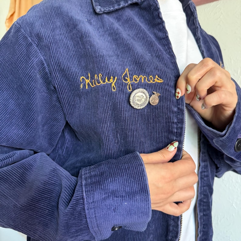 Op de afbeelding: Een marineblauwe corduroy jas met de naam "Milly Jones" geborduurd in goudkleurig garen. De jas heeft een ritssluiting en twee kleine spelden. De manchet van de jas is geknoopt. De jas wordt over een wit shirt gedragen.