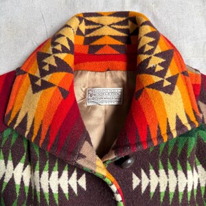 Puede incluir: Primer plano de un abrigo de lana Pendleton con un patr&oacute;n geom&eacute;trico colorido. El cuello presenta un dise&ntilde;o vibrante en tonos naranja, amarillo y marr&oacute;n. El cuerpo del abrigo muestra una mezcla de formas geom&eacute;tricas verdes, blancas y marrones. Se ve un bot&oacute;n.