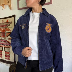 Op de afbeelding: Marineblauwe corduroy jas met ritssluiting. Voorzien van een patch met de tekst "NATIONAL VOCATIONAL AGRICULTURE" en de naam "H. Jones" geborduurd op de voorkant. Inclusief een kleine speld. Een jas in vintage stijl.