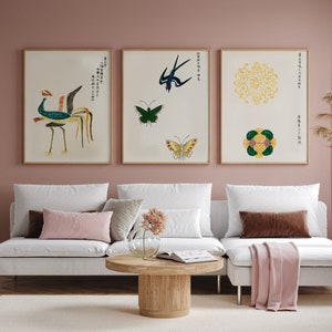May include: Three framed prints with a light pink background. The left print features a colorful bird with a long tail. The middle print features a blue bird and two butterflies. The right print features a yellow mandala and a green and yellow floral design. The prints are hanging on a wall above a white couch with pink and brown throw pillows.