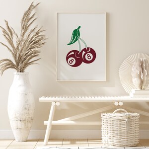 May include: A white framed print with a red and green illustration of two cherries. The cherries are shaped like the number eight billiard balls.
