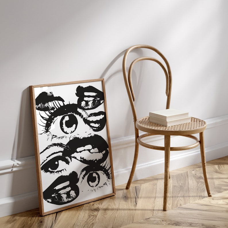 Puede incluir: Impresi&oacute;n en blanco y negro de una cara estilizada con m&uacute;ltiples ojos y labios. La impresi&oacute;n est&aacute; enmarcada en un marco de madera clara y se apoya en una silla de madera clara.