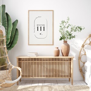K&ouml;nnte beinhalten: Ein gerahmter Druck mit dem Text "INTUITION 111" und dem Satz "Trust Your Gut & Listen to Your Heart". Der Druck h&auml;ngt &uuml;ber einem hellen Holzschrank mit vertikalen Lamellen. Zwei Keramikvasen und eine Pflanze stehen auf dem Schrank.