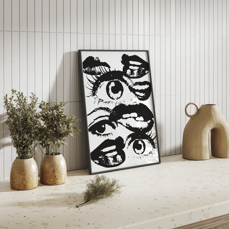 Puede incluir: Impresi&oacute;n enmarcada en blanco y negro de una imagen estilizada de ojos y labios. La impresi&oacute;n est&aacute; colgada en una pared de azulejos blancos sobre un mostrador con dos peque&ntilde;as plantas en maceta y un jarr&oacute;n decorativo.