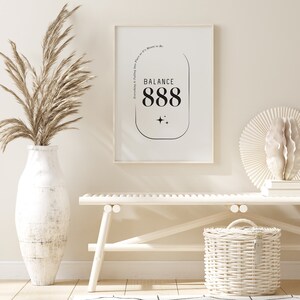 May include: A white framed print with black text that reads "Everything is Falling into Place as it is Meant to Be. Balance 888". The print is hanging on a white wall above a white bench with a white wicker basket.