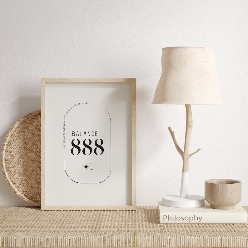 May include: A framed print with a black and white design featuring the text "BALANCE 888" and the phrase "Everything is falling into place as it's meant to be." The print is on a wooden table with a lamp and a book titled "Philosophy".