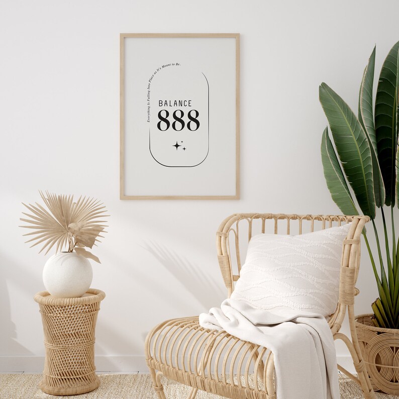 May include: A framed print with a white background and black text that reads "Everything is Falling into Place. It's a Moment to Be... Balance 888". The print is hanging on a white wall above a wicker chair with a white throw blanket and a white pillow.