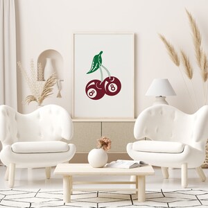 May include: A white framed print with a red and green illustration of two cherries. The cherries are shaped like the number eight billiard balls. The print is hanging on a white wall above a white couch.