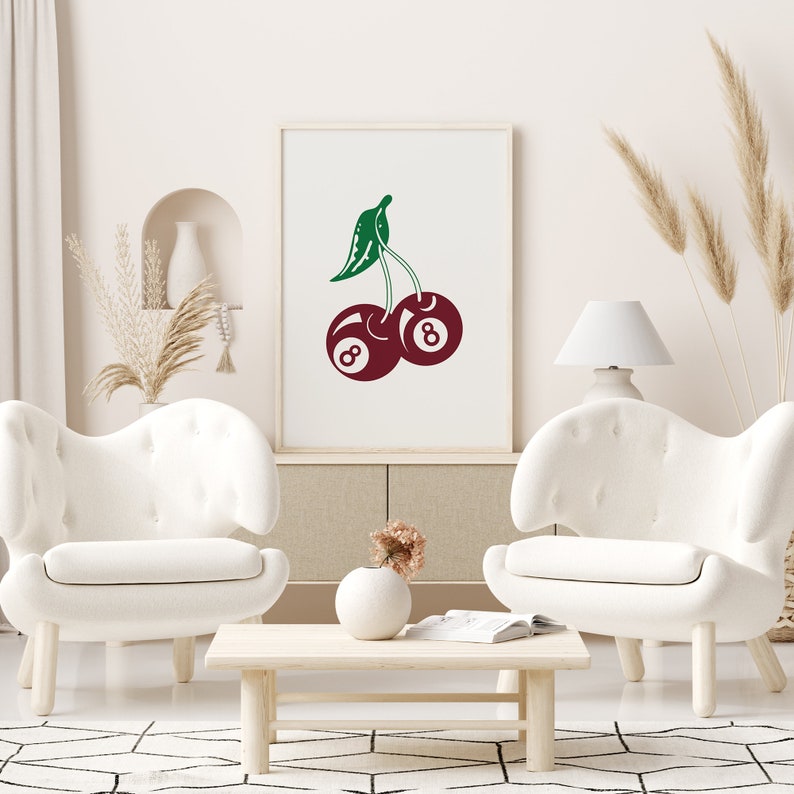 May include: A white framed print with a red and green illustration of two cherries. The cherries are shaped like the number eight billiard balls. The print is hanging on a white wall above a white couch.